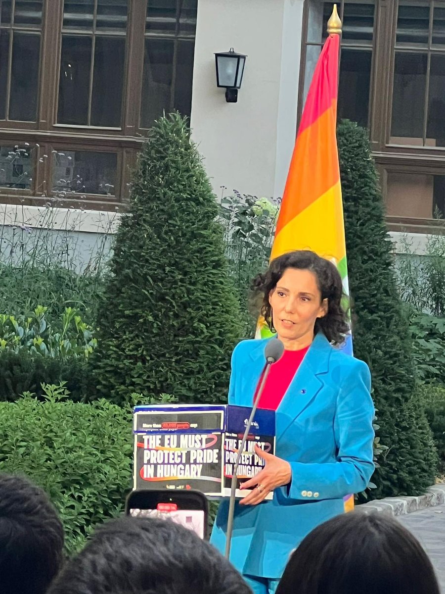 Commissioner Hadja Lahbib speaks at an outdoor podium in front of greenery, with a rainbow Pride flag behind her. She holds a box containing over 40000 signatures and wrapped up in a sign reading “The EU must protect Pride in Hungary” while addressing a crowd, with phones raised to record the moment.